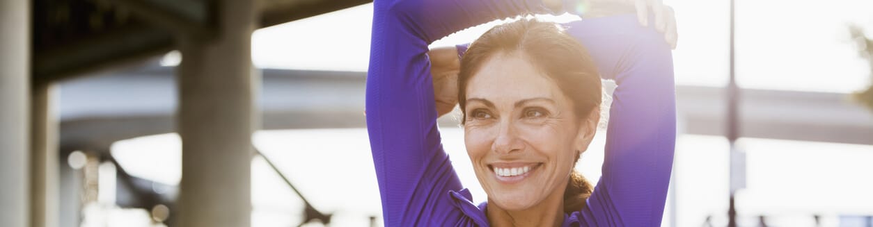 Mulher madura, fazendo uma pausa na corrida para esticar os braços. Ela está ao ar livre em uma passarela de pedestres à beira-mar. É um dia claro e ensolarado. Ela está sorrindo, desviando o olhar da câmera.