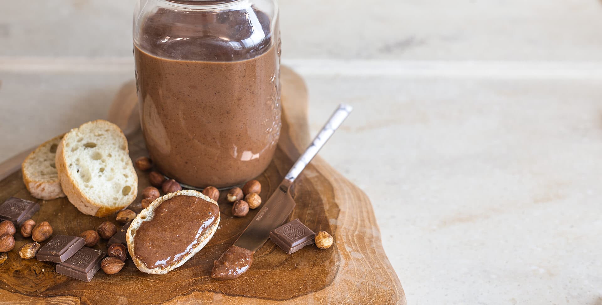 Pote de creme de avelã sem açúcar sob uma tábua de madeira, com pão e avelãs espalhadas