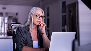 mulher madura, de óculos de grau, em frente ao computador. Ela está concentrada olhando para a tela