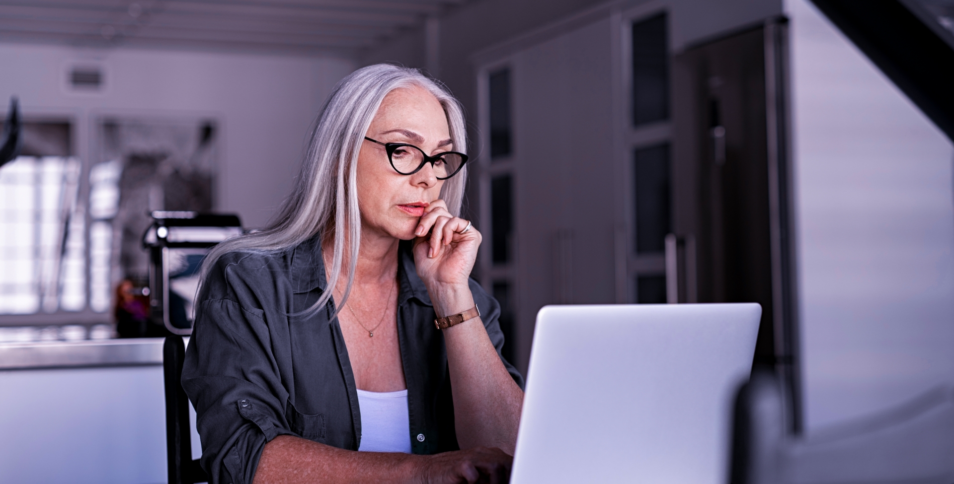 mulher madura, de óculos de grau, em frente ao computador. Ela está concentrada olhando para a tela