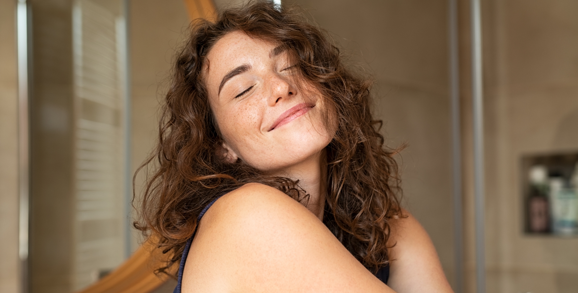 Mulher com os cabelos ondulados está de olhos fechados e sorrindo serenamente enquanto se abraça.